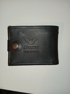 Svart plånbok från Alanwar - Snygg svart plånbok från Alanwar, tillverkad i Syrien. Plånboken är gjord i konstläder och har ett klassiskt vikbart format med knappstängning. Insidan har flera fack för kort och sedlar. Perfekt för dig som vill ha en stilren och praktisk accessoar.