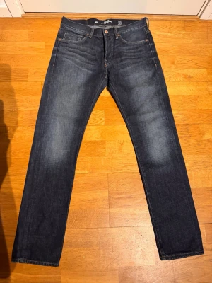Mörkblå raka jeans från Logg - Snygga mörkblå jeans från Logg med klassisk rak passform och fem fickor. Jeansen har subtila slitningar och kontrastsömmar, tillverkade i slitstark denim. Perfekta för en avslappnad och stilren look.