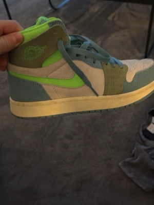 Nike Air Jordan 1 Mid Neon Grön/Blå - Snygga Nike Air Jordan 1 Mid sneakers i beige mocka med detaljer i ljusblått och neonlimegrönt. Klassisk hög siluett, perforerad tåbox och ikonisk Swoosh i neon. Skön vadderad plös och snörning i matchande färg. Perfekta för dig som vill sticka ut.