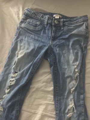 Blå slitna jeans Gina Tricot - Säljer ett par blå jeans från Gina Tricot med slitningar och hål på benen. Jeansen har fem fickor, knappgylf och dekorativa sömmar på bakfickorna. Perfekta för en avslappnad och trendig look.