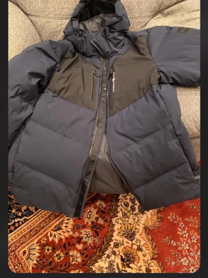 Mörkblå pufferjacka från Sail Racing XXL - Snygg mörkblå pufferjacka från Sail Racing med svarta detaljer på bröstet och huva. Jackan har dragkedja framtill, två bröstfickor med dragkedja och en hög krage. Perfekt för kalla vinterdagar tack vare det tjocka materialet. Storlek XXL