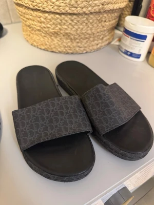 Tofflor Dior - Snygga svarta slides från Dior med klassiskt monogrammönster på remmen. Sandalerna har en bred textilrem och en bekväm sula i syntetmaterial. Perfekta för en avslappnad och trendig look.