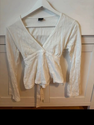 Vit knyttop från Gina Tricot - Säljer en vit omlottopp från Gina Tricot i storlek XS. Toppen har långa ärmar, v-ringning och knytband i ryggen. Materialet är mjukt och lätt, perfekt för en fräsch och enkel look. 