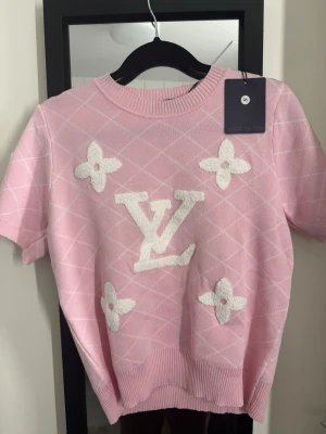 Rosa tröja, kort ärm - Supersöt rosa tröja med korta ärmar och ribbade muddar. Tröjan har ett diskret rutmönster och blommor framtill. Perfekt för dig som vill sticka ut med en lyxig och trendig look.