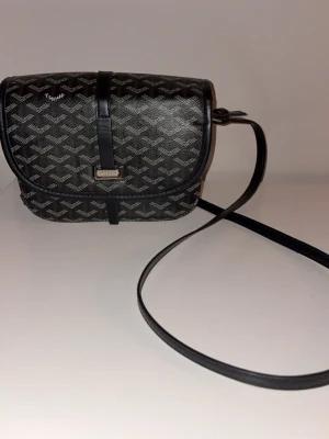 Svart axelväska från Leopard - Snygg svart axelväska från Leopard med klassiskt mönster i grått och vitt. Väskan har rundad form, lock med spänne och lång justerbar axelrem. Tillverkad i konstläder med metall-detaljer framtill. Perfekt för dig som vill ha en stilren och trendig väska.