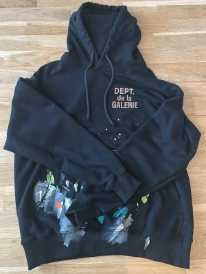 Svart hoodie Dept. de la Galerie - Svart hoodie med tryck 'DEPT. de la GALERIE' på bröstet och färgstänk i blått, grönt, vitt och rött på nedre delen. Klassisk huva med dragsko och ribbade muddar. Perfekt för dig som gillar streetwear och unika detaljer.
