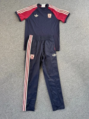 Arsenal x Adidas träningsset marinblå - Snyggt träningsset från Adidas och Arsenal i marinblått med vinröda detaljer. T-shirten har vita och röda ränder på axlarna och Arsenal-logga på bröstet. Byxorna har matchande ränder längs benen och elastisk midja. Perfekt för fotboll eller träning.