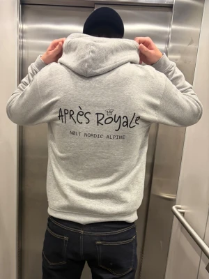 Grå hoodie Après Royale - Snygg grå hoodie från Nolt Nordic Alpine med stor svart text 'Après Royale' och en liten krona på ryggen. Hoodien har en klassisk huva och ribbade muddar. Perfekt för dig som gillar streetwear och vill ha en chill look.