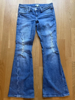 Blå bootcut jeans med slitningar - Snygga blå bootcut jeans med slitna detaljer på båda benen och klassisk femficksdesign. Jeansen har låg midja och utställda ben. Jeansen har en liten defekt på ena lårbenet(se sista bilden)💗