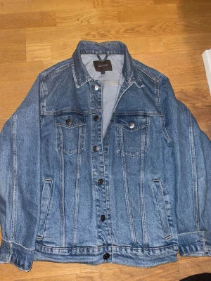 Blå jeansjacka från Dressmann - Klassisk blå jeansjacka från Dressmann med knappar framtill, två bröstfickor med lock och knapp samt sidofickor. Jackan har en rak passform och tydliga sömdetaljer som ger en tidlös look.