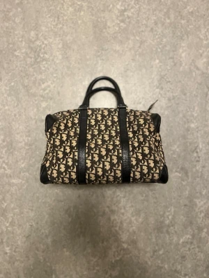 Dior bowling cloth handbag Vintage - Säljer en snygg handväska från Dior i beige och svart canvas med klassiskt monogrammönster. Väskan har svarta läderdetaljer och dubbla handtag. Perfekt för dig som gillar lyxiga accessoarer med ikonisk design. Skriv gärna om du har funderingar eller något annat😁