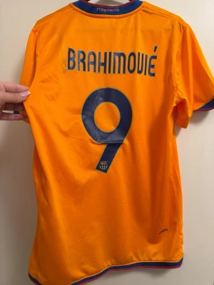 FC Barcelona Ibrahimović matchtröja - Säljer en orange FC Barcelona matchtröja med blå detaljer, nummer 9 och Ibrahimović på ryggen. Tröjan har klubbnamnet broderat i nacken och är tillverkad i lätt och ventilerande polyester, perfekt för fotbollsträning eller som supporterplagg.