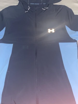 Blå vindjacka från Under Armour - Snygg marinblå vindjacka från Under Armour med ljusblå paneler på sidorna och ärmarna. Jackan har huva med snörning, dragkedja framtill och logga på bröstet. Perfekt för dig som vill ha en sportig look.
