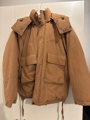 Brun pufferjacka från BOG Jeans - Säljer en brun pufferjacka från BOG Jeans med stor huva och två rejäla fickor framtill. Jackan har dragsko i nederkant och hög krage med tryckknappar. Perfekt oversized look och riktigt skön för kalla vinterdagar.