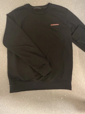 Svart sweatshirt från Prada - Säljer en svart sweatshirt från Prada med rund halsringning och diskret logga på bröstet i rött och vitt. Tröjan har en klassisk passform med ribbade muddar vid ärmslut och nederkant. Perfekt för en stilren och trendig look.
