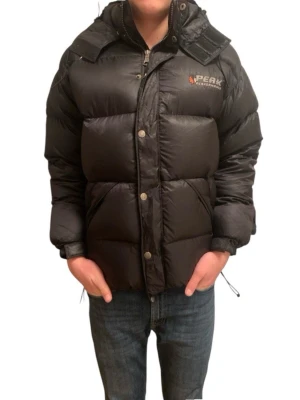 Svart dunjacka Peak Performance - Svart dunjacka från Peak Performance med hög krage, dragkedja och tryckknappar. Jackan har en klassisk puffer-modell och logga på bröstet. Perfekt för kalla vinterdagar och riktigt varm tack vare dunfyllningen. Modellen är 177cm