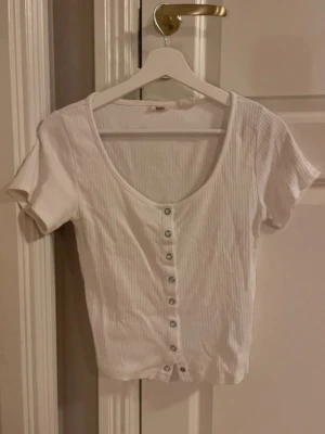 Vit ribbad topp från Levi's - Supersöt vit ribbad topp från Levi's med korta ärmar och djup v-ringning. Toppen har knappar hela vägen framtill och är i mjuk bomull som sitter snyggt och bekvämt. Perfekt till jeans eller kjol för en clean och fräsch look.