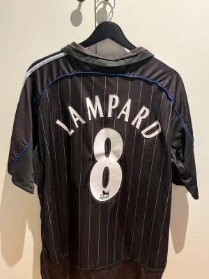 Chelsea tröja svart - Säljer en svart Chelsea FC matchtröja från Adidas med Lampard och nummer 8 på ryggen. Tröjan har vita och blå detaljer, tunna vertikala ränder och klubbmärke på bröstet. Samsung Mobile som sponsor framtill. Perfekt för fotbollsfans!