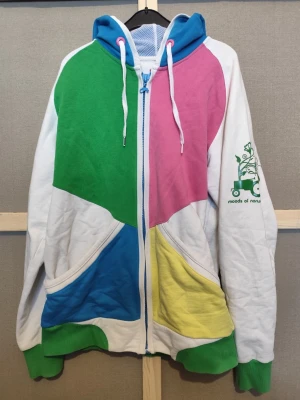 Färgglad hoodie från Moods of Norway - Säljer en unik hoodie från Moods of Norway med färgblock i grönt, rosa, blått och gult. Dragkedja framtill, två fickor och vit bas. Grön logga med traktor och blommor på ärmen. Huva med blå insida och vita snören. Perfekt för dig som gillar statement-plagg!