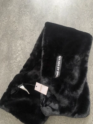 Svart fuskpäls halsduk från Bershka - Snygg svart halsduk i mjuk fuskpäls från Bershka med texten 'COME AS YOU ARE' broderad på ena sidan. Perfekt för att ge din outfit en cool och trendig touch. Lång och rektangulär modell som känns lyxig och varm.