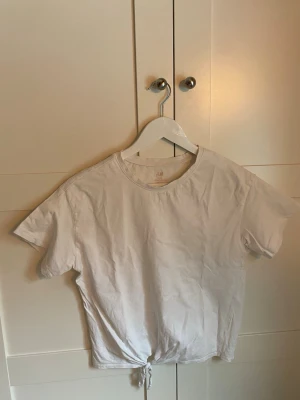 Vit knyt-topp från H&M - Säljer en enkel och fräsch vit topp från H&M med korta ärmar och knyt i nederkanten. Knuten går att ta upp. Toppen har en rund halsringning och är gjord i mjuk bomull, perfekt till jeans eller kjol.                                                   Texten i nacken är bortskavd, därav svårt att läsa av storlek. 