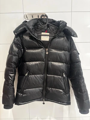 Svart dunjacka från Moncler - Snygg svart dunjacka från Moncler i modellen Maya. Jackan har en glansig finish, huva och två dragkedjeförsedda fickor. Jackan har breda kanaler och en hög krage som skyddar mot kyla. Perfekt för kalla vinterdagar och riktigt stilren look. 