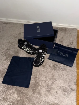 Svarta Dior B30 sneakers strl 46 - Säljer ett par svarta Dior B30 sneakers med stora CD-loggor på sidorna och vita detaljer. Skorna har snörning, platt sula och rund tå. Materialet är en mix av mesh och syntet, vilket ger en sportig och lyxig vibe. Kommer med originalkartong och dustbag.