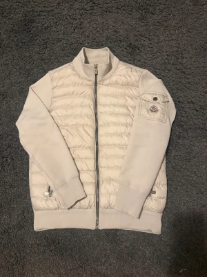 Moncler cardigan - Snygg cremevit cardigan från Moncler med quiltad front och stickade ärmar. Jackan har dragkedja framtill, en ficka med knapp på ärmen och Moncler-logga. Perfekt för dig som gillar stilren design och exklusiva märken.