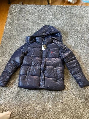 Mörkblå pufferjacka Polo Ralph Lauren - Säljer en mörkblå pufferjacka från Polo Ralph Lauren med huva och dragkedja. Jackan har det klassiska röda Polo-logot på bröstet, flera fickor och en glansig finish. Perfekt för kalla dagar och riktigt snygg till streetwear-stilen. Pris kan diskuteras säljer den för billigt för övre fickan är sönder