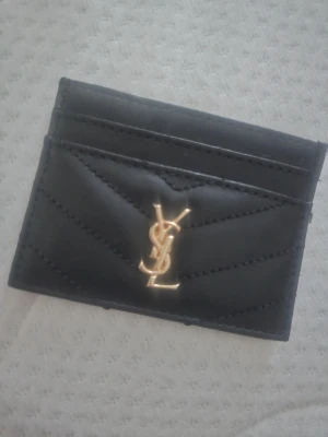 Svart  korthållare i skinn - Säljer en svart korthållare i mjukt skinn med quiltade sömmar och ikonisk guldfärgad logga framtill. Smidig och stilren design med flera kortfack, perfekt för dig som vill ha något både classy och praktiskt i väskan.