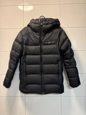 Svart dunjacka Sail Racing med huva - Svart dunjacka från Sail Racing med huva och diskret logga på bröstet. Jackan har quiltad design, dragkedja framtill och är fylld med 80/20 dun/fjäder (600 fill power). Perfekt för kalla dagar. Jackan är i nyskick och har inga defekter, alla märken är kvar och inga hål. Man kan anpassa storleken på jackan de finns spänningar vid midjan och handlederna. Passar någon som är mellan 168-180. Nypris 3000kr säljer den för halva priset alltså 1499 .Riktigt fin jacka!