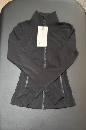 Svart Define jacka från Lululemon storlek 2 - Snygg svart Define jacka från Lululemon med figurnära passform och hög krage. Jackan har dragkedja framtill, långa ärmar med tumhål och är tillverkad i mjukt Nulu™-material (nylon och elastan) som är superstretchigt och bekvämt. Perfekt för träning eller chill.