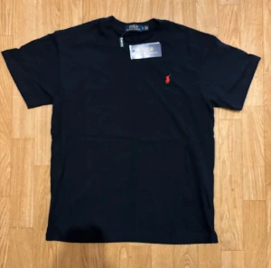 Svart t-shirt från Polo Ralph Lauren - Svart t-shirt från Polo Ralph Lauren med klassisk rund hals och korta ärmar. T-shirten har en röd broderad logga på bröstet och är gjord i mjuk bomull. Perfekt för en clean och stilren look.