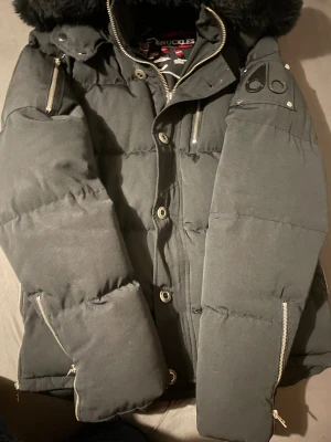 Svart pufferjacka Moose Knuckles - Säljer en svart pufferjacka från Moose Knuckles med dunfyllning och snygga detaljer som dragkedjor på ärmar och bröst, samt logga på axeln. Jackan har hög krage med fuskpäls och knappar framtill. Perfekt för kalla dagar.