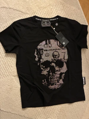 Svart t-shirt med döskalle från Philipp Plein - Cool svart t-shirt från Philipp Plein med glittrande döskallemotiv framtill. Rund hals och korta ärmar. Materialet är en mix av bomull och polyester, vilket gör den mjuk och skön. Märkesdetaljer syns både i nacken och på sidan. Perfekt för dig som gillar edgy stil.