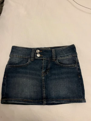 Blå jeanskjol från H&M Divided - Snygg blå jeanskjol från H&M Divided i klassisk femficksmodell med dubbla silverknappar fram och bälteshällor. Kjolen är kort och har en rak passform, perfekt till sneakers eller boots. Materialet är slitstark bomull med kontrastsömmar.