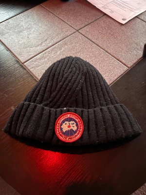 Svart ribbstickad mössa Canada Goose - Svart ribbstickad mössa från Canada Goose med klassisk rund röd och blå patch framtill. Insidan är fodrad med mjuk grå fleece för extra värme. Perfekt för kalla dagar och har en snygg, enkel design som funkar till allt.