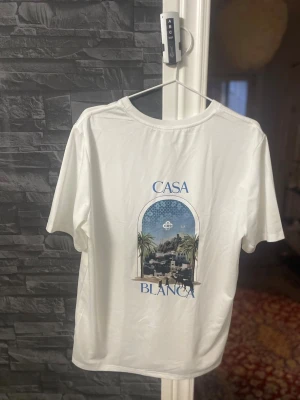 Vit Casa Blanca t-shirt med tryck - Vit t-shirt från Casa Blanca med snyggt tryck av palmer och stadsmotiv både fram och bak. Klassisk rund hals och korta ärmar. Perfekt för dig som gillar stilrena plagg med coola detaljer.
