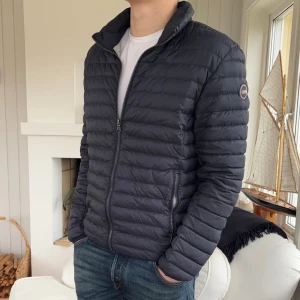 Mörkblå dunjacka från Colmar - Snygg mörkblå dunjacka från Colmar med quiltad design och dragkedja framtill. Jackan har ståkrage, två sidofickor med dragkedja och ett diskret Colmar-märke på ärmen. Perfekt för kyliga dagar och har en clean, modern look. Storlek 52