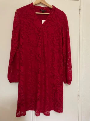 Röd spetsklänning från Mywear Woman - Röd kortklänning i spets med blommigt mönster från Mywear Woman. Klänningen har v-ringning, långa ärmar och är fodrad undertill. Perfekt för dig som vill sticka ut med en färgstark look. Säljes i storlek S.