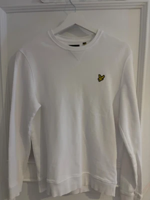 Vit sweatshirt från Lyle & Scott - Vit sweatshirt från Lyle & Scott med klassisk rund hals och långärmad passform. Tröjan har den ikoniska gula fågeln broderad på bröstet och är tillverkad i 100% bomull för en skön och mjuk känsla. Perfekt för en clean och stilren look.