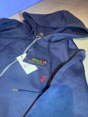 Marinblå hoodie från Polo Ralph Lauren - Snygg marinblå hoodie från Polo Ralph Lauren med dragkedja och huva. Tröjan har en klassisk röd logobrodyr på bröstet, snörning vid huvan och praktiska fickor framtill. Tillverkad i mjuk bomullsmix som är skön att bära. Perfekt för chill dagar.