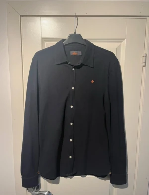 Svart långärmad skjorta från Morris - (Julklapps tips🎄🎁)Svart skjorta från Morris med klassisk krage, vita knappar och lång ärm. Broderad orange logga på bröstet och diskret Morris-detalj vid nederkanten. Skjortan är i bomull och har en clean, stilren look som funkar till många outfits.