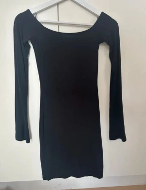 Svart långärmad kortklänning. Gina trixig - En svart, figurnära kortklänning med lång ärm och bred, rundad halsringning. Enkel och stilren design som passar dig som gillar minimalistiskt mode. Mjuk och stretchig känsla i materialet för en bekväm passform.