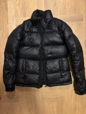 Svart pufferjacka från Peak Performance XS - Svart pufferjacka från Peak Performance i storlek XS. Jackan har hög krage, två sidofickor med dragkedja och quiltad design. Materialet är syntet och dun, perfekt för kalla dagar. Snygg och enkel stil som funkar till många outfits. Har en liten lagning på ärmen som knappt syns men tog kort på den. Jag är 164 cm lång