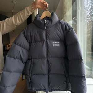 Svart pufferjacka från Axel Arigato - Svart pufferjacka från Axel Arigato med hög krage och diskret logga på bröstet. Jackan har dragkedja framtill, två sidofickor och är tillverkad av återvunnen polyester och dun. Perfekt för kalla dagar och har en clean, modern look.