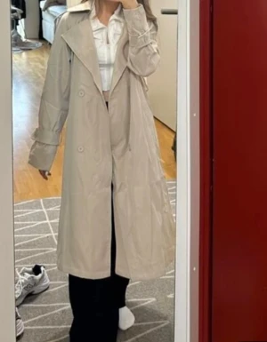 Beige trenchcoat med bälte - Snygg beige trenchcoat med klassisk krage och breda slag. Jackan har långa ärmar med dekorativa remmar vid handlederna och knyts med bälte i midjan. Dubbelknäppt framtill och rak, loose passform som ger en chill vibe. Perfekt för dig som vill ha en stilren och tidlös look.