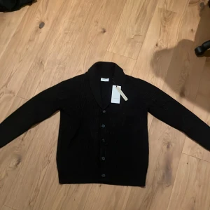 Svart ribbad kofta från Lindbergh - Svart kofta från Lindbergh med ribbad struktur och klassisk krage. Koftan har knäppning framtill med svarta knappar och långa ärmar. Perfekt för lager-på-lager och stilren look.