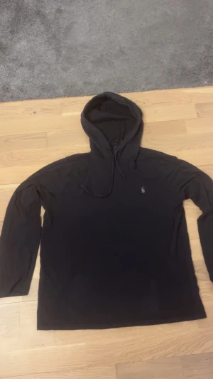 Svart hoodie från Polo Ralph Lauren - Svart hoodie från Polo Ralph Lauren med klassisk luva. Liten broderad logga på bröstet i vitt. Mjukt bomullsmaterial och långärmad modell, perfekt för chill dagar. Snygg och enkel design som funkar till det mesta. OBS defekt på andra bilden! 