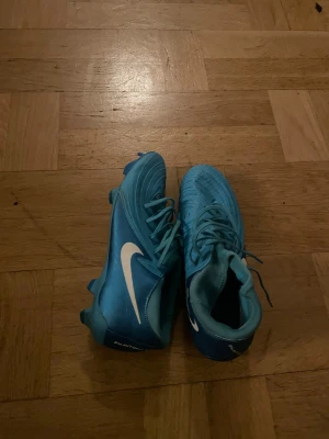 Nike Phantom blå fotbollsskor - Säljer ett par Nike Phantom fotbollsskor i en snygg ljusblå färg med vit swoosh på sidan. Skorna har snörning och är designade för gräsplan med dobbar under sulan. Materialet är syntet och skon har en rund tå för bra bollkontroll.